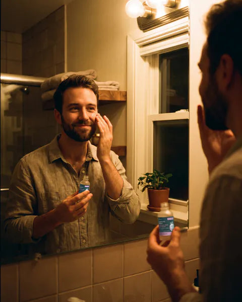 A man at the bathroom mirror presses a few drops of serum onto his cheek, holding a bottle of Hyaluronic Serum.