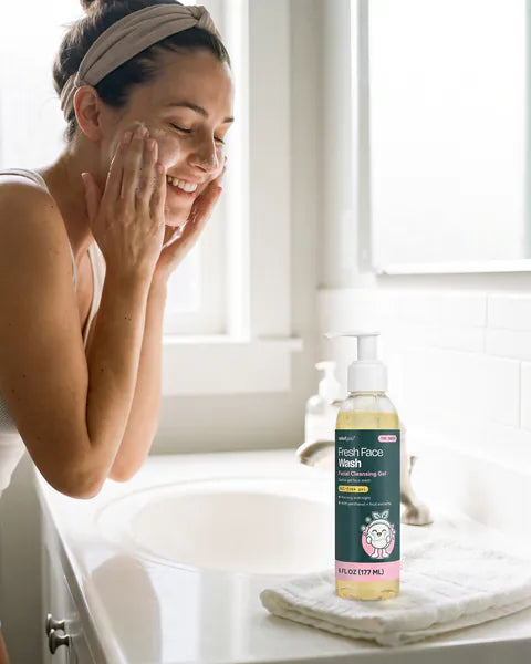 A woman washes her face at a bright bathroom sink in the morning, a bottle of Fresh Face Wash beside the basin.