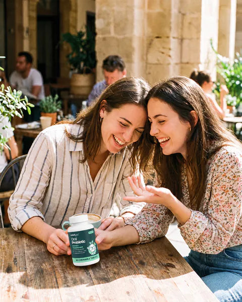 Two friends lean in close, laughing at a café table, with a bottle of Oral Probiotic in front of them.