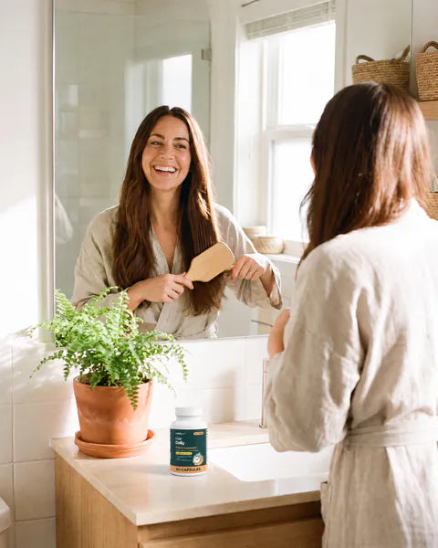 A woman brushes her hair at the bathroom mirror, a bottle of Hair Daily on the counter.