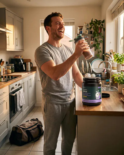 A man laughs as he shakes a shaker bottle in his kitchen, a tub of Protein Refuel Chocolate on the counter.