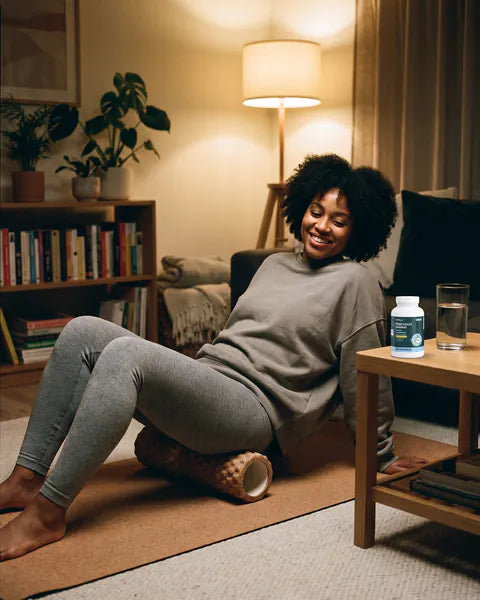 A woman uses a foam roller on a mat in her living room in the evening, a bottle of Magnesium Unwind on the side table.