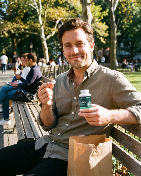A man on a park bench after lunch holds up a chewable tablet and a bottle of Oral Probiotic.