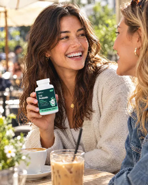 A woman laughs with a friend at a café terrace, holding a bottle of Oral Probiotic.