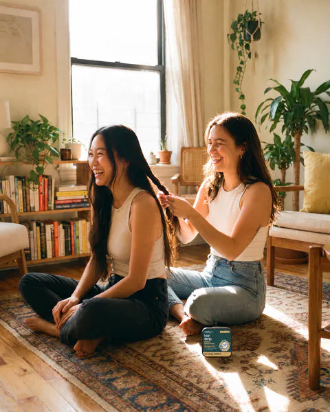 Two friends sit on a living room rug, one braiding the other’s hair, a tin of Hair Strip beside them.