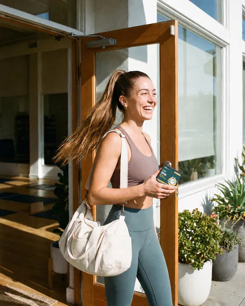 A woman with a high ponytail walks out of a yoga studio, a tin of Hair Strip in her hand.