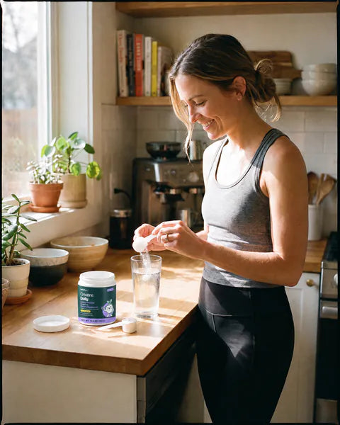 A woman in running clothes stirs a scoop of powder into a glass of water, a tub of Creatine Daily open on the counter.